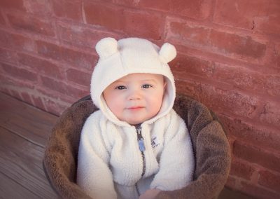 First and Forever Photography, Iveta Dietz, photography by Iveta Dietz, baby in a bucket, white hoodie, bear hoodie, baby, baby boy, best newborn photographer, best baby photographer, best St. Charles photographer, St. Louis photographer, posed baby, sitting baby, baby's first year, safe posing, safety first, comfortable posing, cute baby, happy baby, red brick background, bronw bucket, cute look, big eyes,